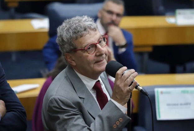 A foto mostra Nabil Bonduki em um ambiente formal, como uma reunião ou conferência. Ele é um homem mais velho, com cabelos grisalhos e cacheados, usa óculos de armação vermelha, terno cinza claro, camisa branca e gravata vermelha. Está segurando um microfone enquanto fala, transmitindo a ideia de participação ativa em um debate ou apresentação. Ao fundo, há outras pessoas sentadas, mesas com papéis e placas de identificação, reforçando o contexto institucional da cena.