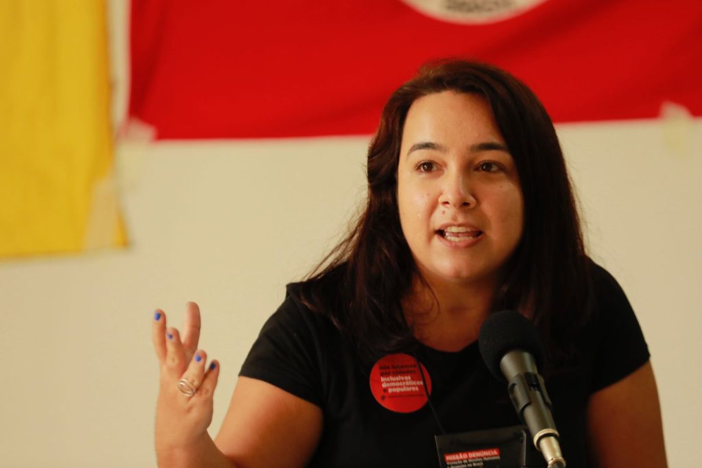Na foto, vemos Raquel Lurdemir falando ao microfone, em um momento que transmite engajamento e firmeza. Ela veste uma camisa preta com dois broches vermelhos, um deles trazendo a frase “nós lutamos por inclusivas democráticas populares” e o outro parcialmente visível com o texto “missão denúncia”. Ao fundo, aparecem duas bandeiras penduradas na parede — uma vermelha e outra amarela — reforçando o ambiente de mobilização social e política. A cena sugere participação ativa em um espaço de discussão ou militância.