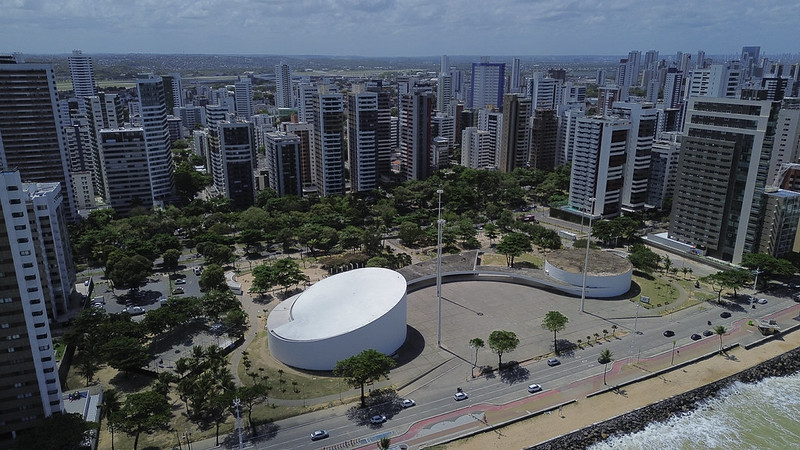 Vista aérea do Parque Dona Lindu, no bairro de Boa Viagem, Recife. No centro da imagem, o edifício oval branco do auditório do parque se destaca em meio a uma área arborizada com gramados e coqueiros. Ao redor do parque, avenidas com carros em movimento e, ao fundo, uma densa fileira de prédios residenciais de vários andares. À direita, a orla marítima com pedras de contenção e o mar.
