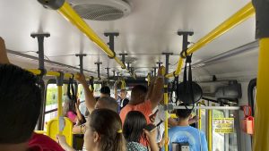Interior de ônibus lotado, passageiros em pé segurando barras amarelas.