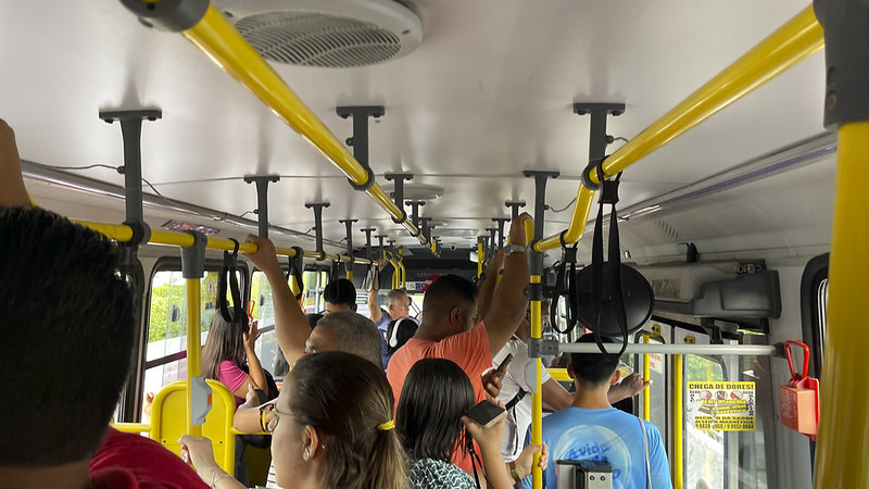 Interior de ônibus lotado, passageiros em pé segurando barras amarelas.