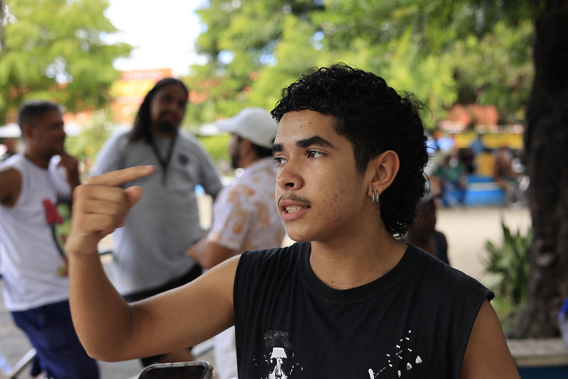 Jovem de cabelo curto e camiseta preta gesticula em conversa, rodeado por pessoas em área pública com árvores.