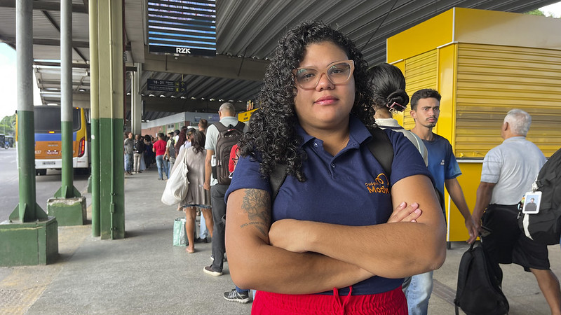 Mulher jovem de óculos e camisa azul, está de braços cruzados em uma fila de terminal de ônibus, com pessoas ao fundo.