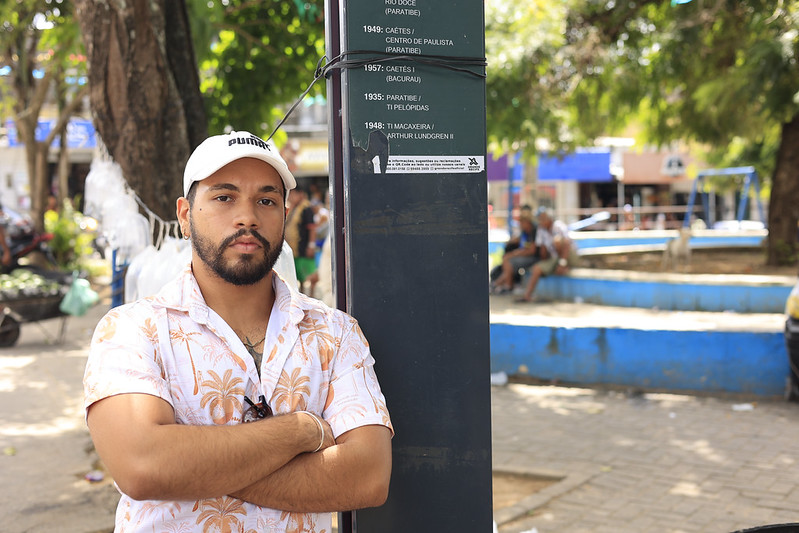 Homem jovem de boné e camisa florida ao lado de uma parada de ônibus com textos de rotas antigas, em praça arborizada.
