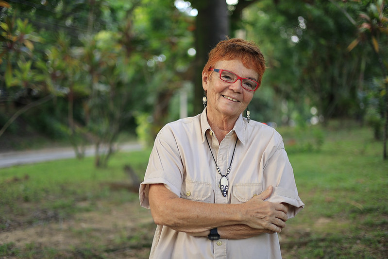 Letícia Lins aparece em um ambiente verde e tranquilo, provavelmente um parque ou jardim, com árvores e folhagem ao fundo. Ela está em pé, ligeiramente voltada para a câmera, com os braços cruzados de forma relaxada sobre o peito. Letícia tem pele clara, cabelo curto e ruivo, usa óculos de armação vermelha e brincos pequenos. Veste uma camisa bege de mangas curtas e um colar com pingente escuro. Seu sorriso é acolhedor e transmite simpatia e serenidade. A luz natural ilumina suavemente seu rosto, sugerindo que a foto foi tirada durante o dia.
