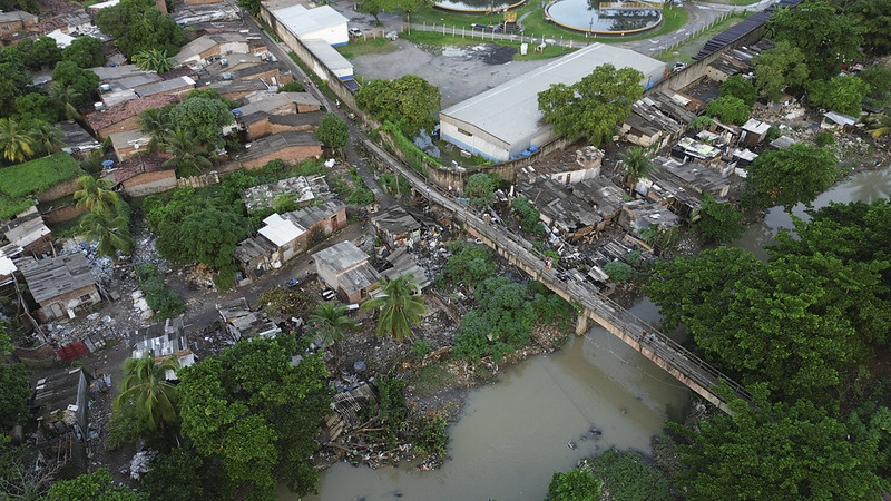 A imagem mostra uma vista aérea de uma área urbana densamente ocupada, com casas simples e construções improvisadas próximas a um rio de águas turvas. Um estreito passarela de madeira atravessa o rio, ligando os dois lados da comunidade. As moradias estão muito próximas umas das outras, e há vegetação misturada com entulho nas margens. No canto superior, vê-se um galpão e uma área pavimentada, sugerindo uma zona industrial ou comercial próxima.