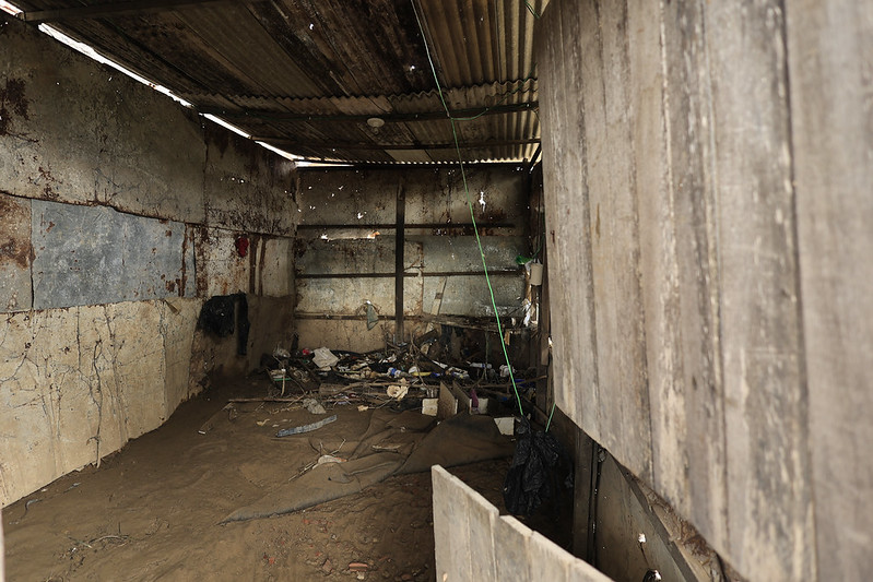 A imagem mostra o interior de uma casa muito simples e deteriorada, construída com chapas metálicas enferrujadas e tábuas de madeira. O teto é de zinco corroído, com furos por onde entra luz natural. O chão é de terra batida, coberto por entulhos e objetos quebrados, como pedaços de madeira e plásticos. Há também um fio verde pendurado do teto, sugerindo improvisação elétrica. O ambiente parece abandonado ou em ruínas, sem móveis ou sinais de uso recente.