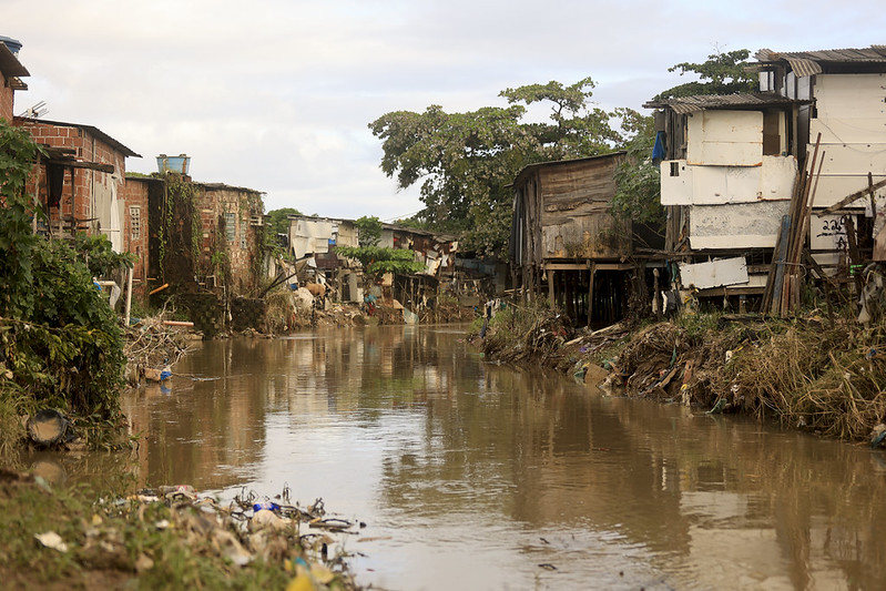 A imagem mostra o rio Beberibe entre Recife e Olinda, cercado por casas simples e muito próximas à margem. As construções são feitas de madeira, tijolos e chapas metálicas, algumas apoiadas sobre estacas para evitar contato direto com a água. O rio aparece barrento e cheio de resíduos, com vegetação e lixo acumulado nas bordas. Ao fundo, há árvores grandes e o céu parcialmente nublado, sugerindo um dia após chuva.