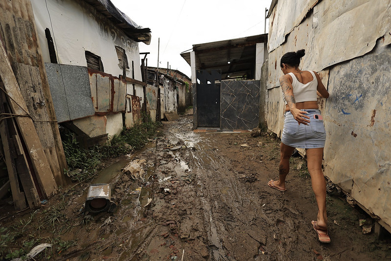 A imagem mostra uma rua estreita e enlameada em uma área de moradias simples. As casas são feitas de materiais reaproveitados, como tábuas, chapas metálicas e pedaços de madeira, com sinais de desgaste e ferrugem. No centro, uma mulher caminha com cuidado pelo chão escorregadio, vestindo blusa branca e shorts jeans, enquanto segura a parte de trás da roupa para evitar o barro. O ambiente é úmido, com poças d’água e lixo espalhado, e há vegetação crescendo entre as construções.