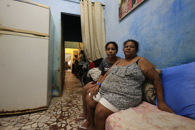 A imagem mostra uma mulher adulta e uma menina sentadas em um sofá simples dentro de uma casa de paredes pintadas de azul. A mulher à direita usa uma blusa estampada e shorts claros, enquanto a jovem ao lado veste uma camiseta escura e shorts. O ambiente é pequeno e modesto, com um refrigerador branco à esquerda e um corredor ao fundo que leva a outro cômodo iluminado em tom amarelado. O chão é coberto por pedaços de cerâmica irregular, e há uma cortina clara separando os espaços.