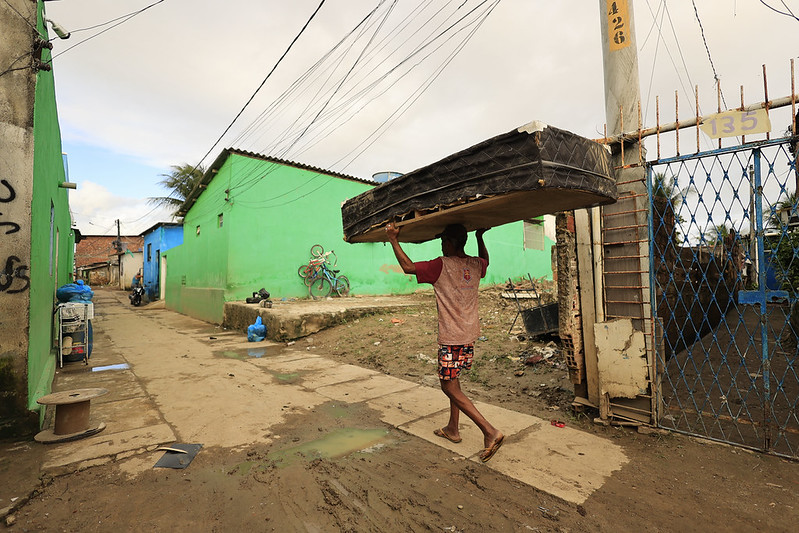 Foto colorida mostra uma rua estreita de terra e cimento em uma comunidade urbana. No centro da imagem, um homem caminha descalço usando bermuda estampada e camiseta rosa enquanto carrega um colchão escuro sobre a cabeça. As casas ao redor têm paredes pintadas em tons fortes de verde e azul, contrastando com o céu nublado. O chão apresenta poças de lama e marcas de desgaste, enquanto fios elétricos atravessam o alto da cena. À direita, há um portão metálico azul e estruturas em construção, reforçando o aspecto simples e cotidiano do local.
