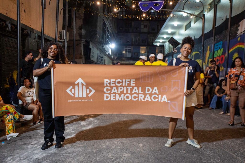 Duas mulheres jovens, negras vestidas com blusas pretas aparecem em uma rua à noite segurando uma faixa laranja com a frase “Recife Capital da Democracia” em letras brancas. No fundo, outras pessoas estão reunidas diante de murais coloridos e decorações festivas. Luzes penduradas e uma grande máscara completam o cenário de celebração comunitária.