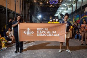 Duas mulheres jovens, negras vestidas com blusas pretas aparecem em uma rua à noite segurando uma faixa laranja com a frase “Recife Capital da Democracia” em letras brancas. No fundo, outras pessoas estão reunidas diante de murais coloridos e decorações festivas. Luzes penduradas e uma grande máscara completam o cenário de celebração comunitária.