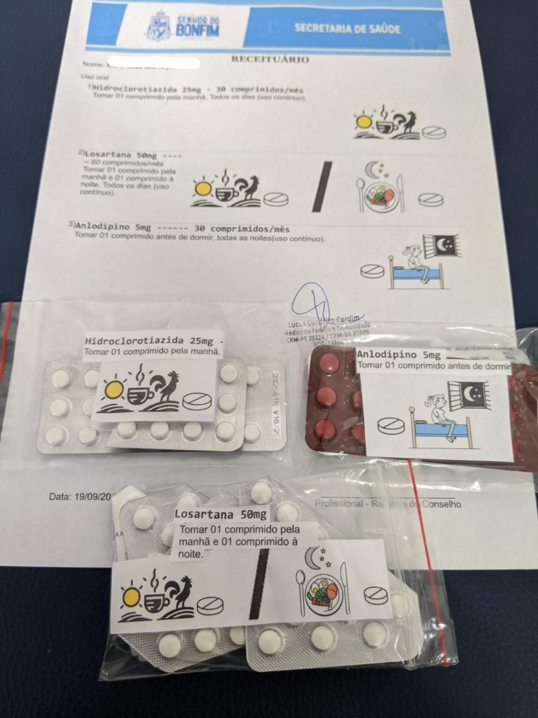 A foto mostra uma receita médica acompanhada das cartelas dos remédios prescritos. O documento é oficial da Secretaria de Saúde de Senhor do Bonfim, assinado por um médico. Sobre a receita estão três cartelas de comprimidos, cada uma com etiquetas impressas que indicam o nome do medicamento, a dosagem e instruções claras de uso. As etiquetas trazem também pequenos ícones (como sol, lua, cama, xícara de café) para ajudar o paciente a identificar o momento certo de tomar cada remédio — de manhã, à noite ou antes de dormir. Em resumo, é uma imagem que combina a prescrição médica com os próprios medicamentos, organizada de forma acessível e visual para facilitar o tratamento diário.