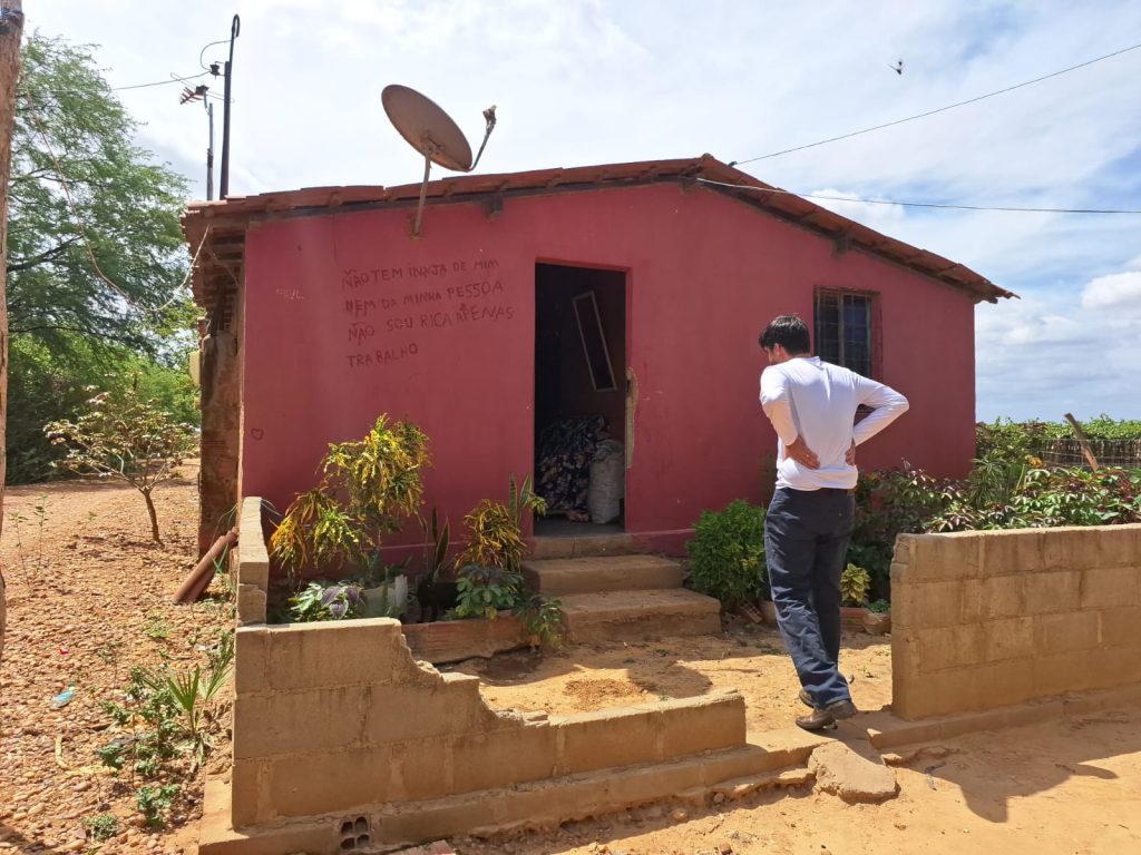 A foto mostra uma pequena casa vermelha, simples, com telhado de telhas e uma antena parabólica. Na frente, há alguns degraus que levam à porta de entrada, cercada por plantas e um muro baixo de tijolos inacabado ou danificado. O chão ao redor é arenoso e seco, com algumas árvores ao fundo. Perto da porta, uma pessoa de camisa branca e calça escura está de pé, olhando para dentro da casa. Na parede, há uma mensagem escrita à mão em português que diz: “Não tem inveja de mim nem da minha pessoa. Não sou rica, apenas trabalho.”