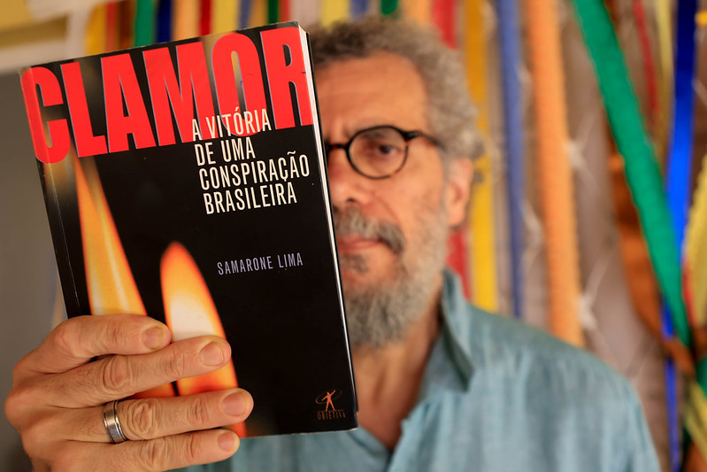 A foto mostra Samarone Lima segurando seu livro Clamor: A vitória de uma conspiração brasileira bem próximo à câmera, de modo que a capa ocupa o primeiro plano. O autor aparece ao fundo, ligeiramente desfocado, com expressão serena e usando óculos e camisa azul. A capa do livro tem letras grandes em vermelho e branco sobre fundo escuro, com duas chamas de vela que sugerem memória e resistência. Atrás dele, há fitas coloridas penduradas, criando um ambiente vibrante e acolhedor.