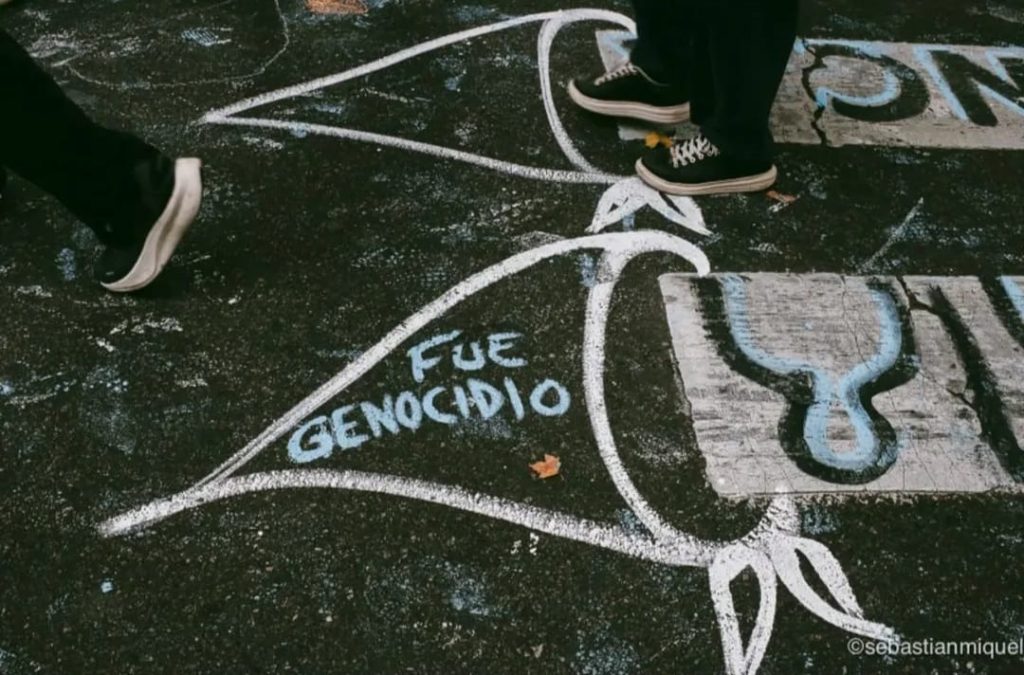A imagem mostra um chão de asfalto coberto por desenhos e palavras feitas com giz branco e azul. No centro, lê-se “FUE GENOCIDIO”, expressão em espanhol que significa “foi genocídio”. O texto está dentro de um contorno que lembra um lenço triangular, símbolo das Mães da Praça de Maio, movimento argentino que denuncia os desaparecimentos durante a ditadura militar. Dois pares de pés com tênis escuros aparecem caminhando sobre ou perto dos desenhos, sugerindo uma cena de rua, possivelmente durante um ato de memória ou protesto. No canto inferior direito, há a assinatura “@sebastianmiquel”.