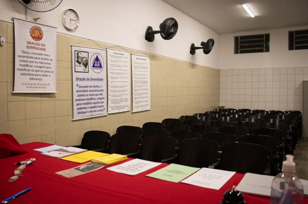 A imagem mostra uma sala de reuniões simples e organizada, com cadeiras pretas alinhadas voltadas para uma parede onde há cartazes com mensagens de apoio e reflexão, incluindo a Oração da Serenidade, os Doze Passos e as Doze Tradições, textos tradicionais de grupos de recuperação como Jogadores Anônimos e Alcoólicos Anônimos. Na frente da sala, uma mesa coberta com toalha vermelha exibe folhetos coloridos, documentos e um frasco de álcool em gel, indicando preparação para uma reunião. O ambiente é iluminado por lâmpadas fluorescentes, possui ventiladores de parede e um relógio.