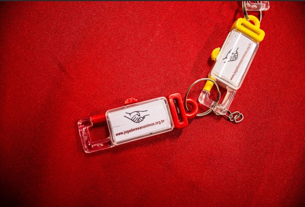 A imagem mostra dois chaveiros plásticos transparentes sobre um tecido vermelho. Cada chaveiro tem detalhes coloridos — um com partes vermelhas e outro com amarelas — e contém um pequeno cartão branco com o desenho de duas mãos se cumprimentando, símbolo de apoio e solidariedade. Abaixo do desenho, lê-se o endereço “www.jogadoresanonimos.org.br”, indicando que são materiais do grupo Jogadores Anônimos, voltado à recuperação de pessoas com dependência em jogos.