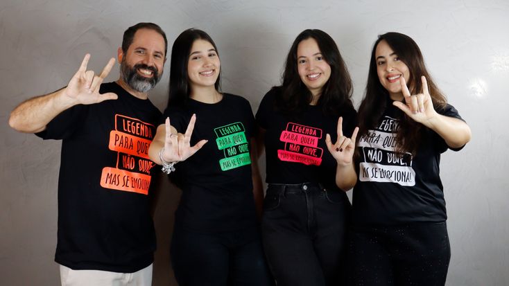 Quatro pessoas posam lado a lado em frente a uma parede clara. Da esquerda para a direita: um homem de barba grisalha e três mulheres de cabelos escuros. Todos vestem camisetas pretas com a frase “Legenda para quem não ouve, mas se emociona!” escrita em diferentes cores (Laranja, verde, rosa e branca). Eles fazem com as mãos o sinal de “eu te amo” em Libras e sorriem para a câmera. Fonte: Acervo Legenda Nacional