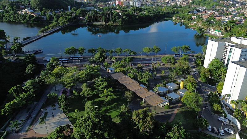 Parque de Apipucos, em Recife, aparece nesta foto aérea como um refúgio verde em meio à cidade: o lago central reluz como um espelho que reflete céu e árvores, enquanto suas margens são cercadas por caminhos sombreados, praças e estruturas acolhedoras que convidam ao descanso; ao lado, uma avenida movimentada lembra o ritmo urbano, e mais adiante os edifícios brancos modernos se erguem em contraste com a natureza, compondo uma paisagem onde se misturam o frescor das copas, o cheiro de grama molhada e a brisa suave do lago com o cenário vibrante da vida recifense.