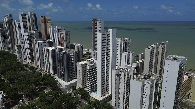 Esta foto mostra uma vista aérea de uma área urbana litorânea, com vários prédios altos e modernos alinhados paralelamente à costa. As construções são predominantemente brancas e cinzas, com variações sutis de cor e design. À esquerda, há uma faixa verde com árvores que acompanha a avenida principal, enquanto ao fundo se estende o mar calmo sob um céu parcialmente nublado.