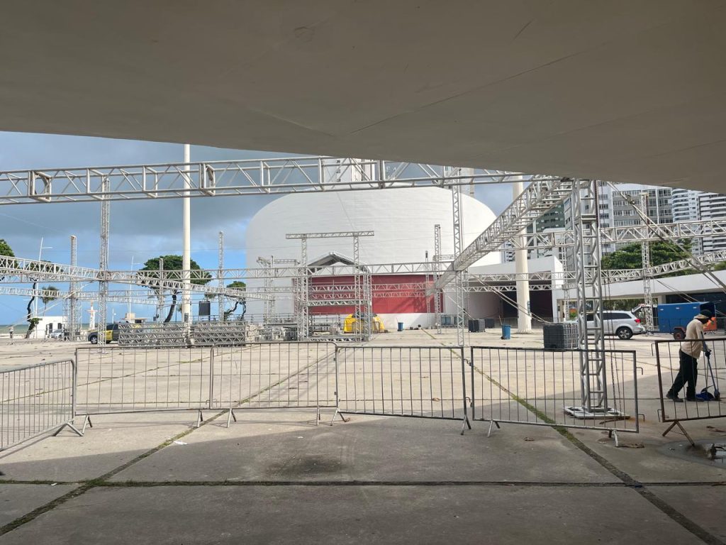 Área externa do Parque Dona Lindu durante montagem de estrutura para evento. No centro da imagem há várias estruturas metálicas prateadas montadas sobre o piso de concreto, cercadas por grades móveis de contenção. Ao fundo aparece o prédio principal do parque, com formato arredondado e fachada branca e vermelha. À esquerda é possível ver o mar e alguns coqueiros; à direita, prédios residenciais altos.