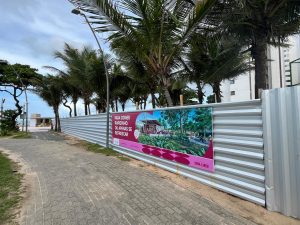 Vista externa do Parque Dona Lindu, em Boa Viagem, Recife. À direita, um tapume metálico branco cerca parte da área do parque. Preso ao tapume há um grande painel colorido com uma imagem ilustrativa e a frase “Para comer rapidinho ou apenas se refrescar”. À esquerda, uma calçada larga de blocos cinza segue em direção à praia, cercada por coqueiros altos e postes de iluminação. O céu está nublado.