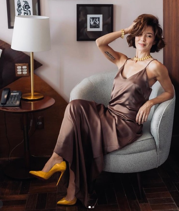 A imagem mostra uma mulher sentada em uma poltrona cinza clara, em um ambiente interno elegante. Ela veste um vestido longo de cetim marrom e sapatos de salto alto amarelos, que criam um contraste marcante. Usa colar e pulseira dourados, e tem o cabelo curto e ondulado, penteado de forma descontraída. Está com uma expressão confiante e um leve sorriso, com uma das mãos apoiada na cabeça. Ao lado, há uma mesa redonda de madeira com um abajur dourado e branco e um telefone preto, compondo um cenário sofisticado e acolhedor.