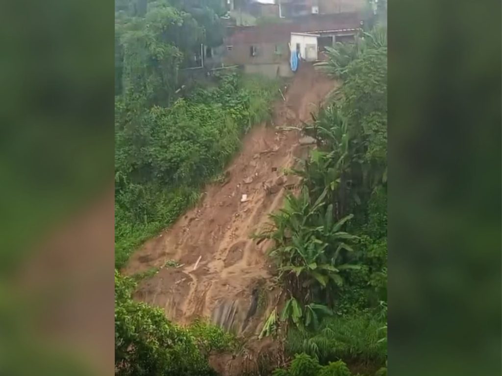 A foto mostra um deslizamento de terra em uma encosta íngreme próxima a casas. Uma grande faixa de solo e lama deslizou, deixando exposta a terra nua. No alto da ladeira, há residências muito próximas da borda, onde o terreno cedeu. Ao redor, a vegetação verde e densa contrasta com a área afetada, reforçando a ideia de um ambiente tropical ou subtropical. É uma cena que evidencia os riscos de construir em terrenos instáveis e sujeitos à erosão.