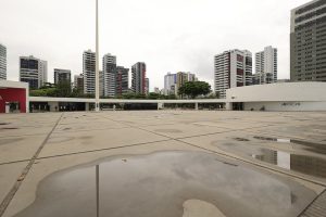 A foto mostra o Parque Dona Lindu, em Recife, com sua grande praça de concreto ainda molhada pela chuva, formando pequenas poças espalhadas pelo chão. Ao fundo, destacam-se vários prédios altos e modernos, criando contraste com o espaço aberto do parque. Em primeiro plano, aparece a construção branca e baixa de linhas simples, parte do complexo cultural projetado por Oscar Niemeyer, onde funciona o Teatro Luiz Mendonça. Algumas pessoas caminham ao longe, e árvores pontuam o cenário com verde, suavizando a paisagem urbana. O céu está nublado, dando um tom mais sóbrio à cena, mas reforçando a atmosfera típica de dias chuvosos na cidade.