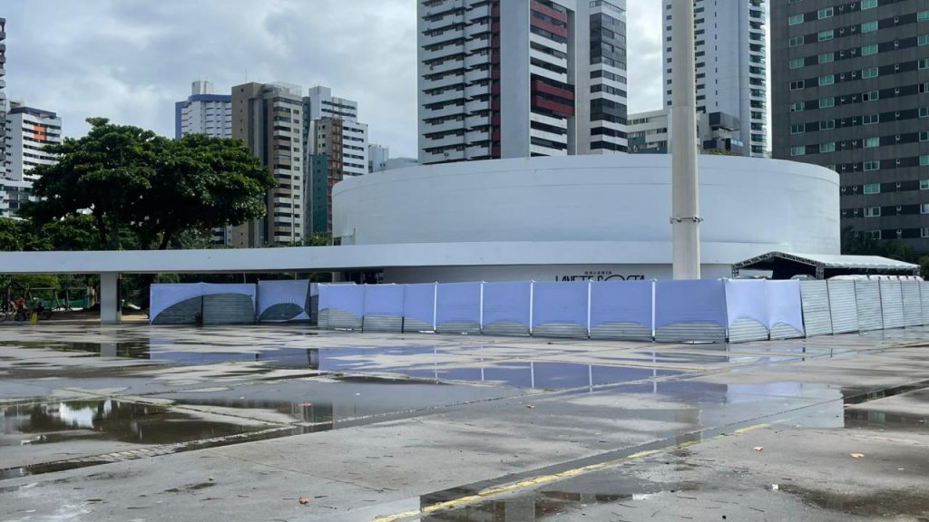 A foto mostra o Parque Dona Lindu, em Recife, com destaque para o prédio circular branco da Galeria Janete Costa. Ele aparece cercado por estruturas metálicas e lonas, indicando obras ou manutenção. Ao fundo, vemos edifícios modernos de diferentes estilos e cores, além de árvores que trazem contraste verde ao cenário urbano. O chão molhado reflete parte das construções e do céu, sugerindo que choveu recentemente.