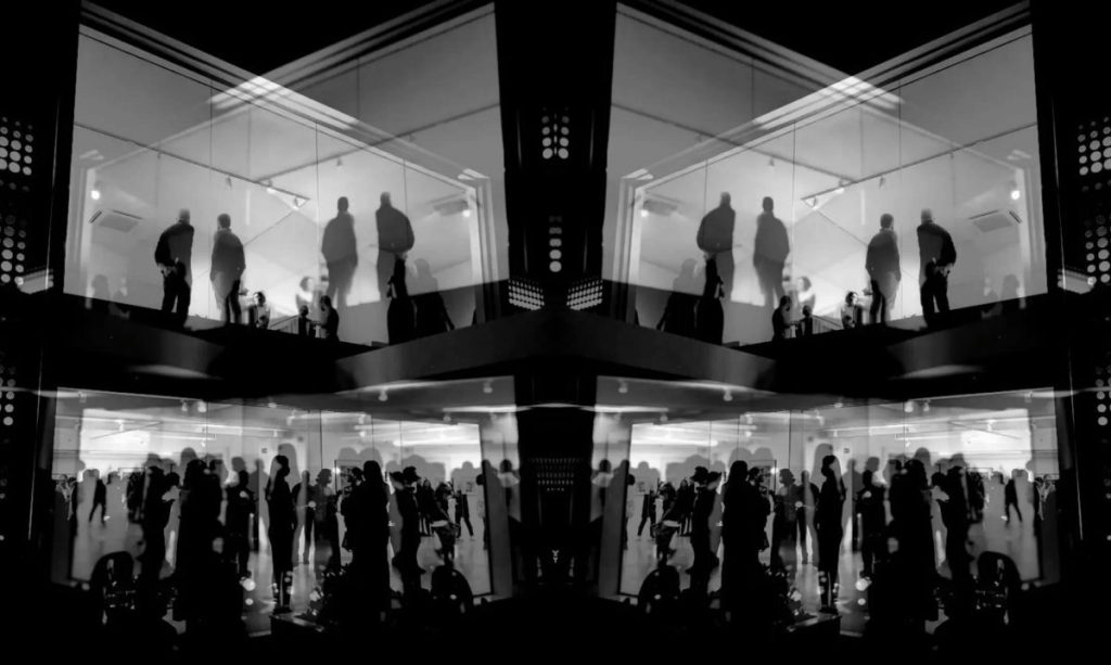 Fotografia em preto e branco de um edifício com fachada de vidro em dois andares. No andar superior, silhuetas de pessoas de pé conversando. No andar inferior, uma multidão maior em movimento. A imagem é espelhada horizontalmente, criando uma composição simétrica.