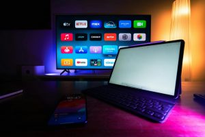 Em um ambiente escuro iluminado por luzes em tons de azul, rosa e amarelo, uma televisão exibe a tela inicial aplicativos da Netflix, Disney+, Prime Video e YouTube. À frente da TV, sobre uma mesa de madeira, há um tablet com teclado acoplado e a tela clara ligada. Um celular aparece desfocado no primeiro plano. À direita, uma luminária alta emite luz amarelada