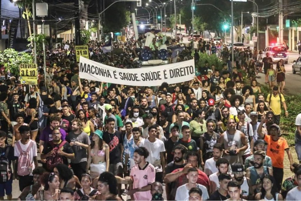 A foto mostra uma grande manifestação noturna em uma avenida urbana, com centenas de pessoas caminhando juntas em clima de mobilização e engajamento. No centro, um grupo segura uma faixa branca com letras pretas que diz “MACONHA É SAÚDE. SAÚDE É DIREITO.”, destacando o caráter político e social do evento. A multidão é diversa, composta por jovens e adultos, alguns sorrindo, outros atentos, e todos participando de forma pacífica. As luzes da rua e dos prédios iluminam o cenário, reforçando a atmosfera vibrante e coletiva. O registro transmite energia, união e reivindicação por direitos e políticas de saúde relacionadas ao uso da cannabis.
