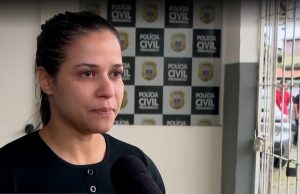 A fotografia mostra Aline Melo em um ambiente institucional, aparentemente uma delegacia da Polícia Civil de Pernambuco — o painel ao fundo confirma isso com o brasão e o nome da corporação repetidos. Ela está sendo entrevistada, já que há um microfone direcionado a ela. Aline parece emocionada, com expressão séria e olhos marejados, sugerindo que fala sobre um assunto delicado ou pessoal. A iluminação é natural e o enquadramento foca em seu rosto e parte superior do corpo.