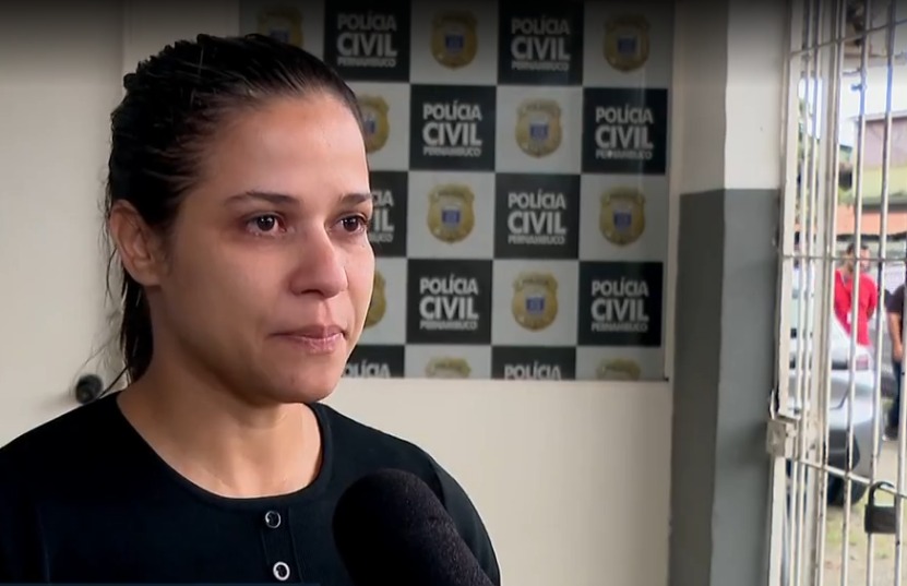 A fotografia mostra Aline Melo em um ambiente institucional, aparentemente uma delegacia da Polícia Civil de Pernambuco — o painel ao fundo confirma isso com o brasão e o nome da corporação repetidos. Ela está sendo entrevistada, já que há um microfone direcionado a ela. Aline parece emocionada, com expressão séria e olhos marejados, sugerindo que fala sobre um assunto delicado ou pessoal. A iluminação é natural e o enquadramento foca em seu rosto e parte superior do corpo.