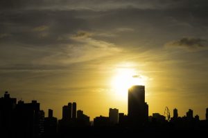 A foto mostra o céu ao entardecer sobre uma cidade. O sol está baixo, quase se pondo, e sua luz dourada ilumina as nuvens e cria um brilho intenso atrás dos prédios. As construções aparecem em silhueta — escuras e sem detalhes — formando uma linha irregular contra o fundo luminoso. O contraste entre o brilho do sol e as sombras dos edifícios dá uma sensação de calma e beleza urbana.
