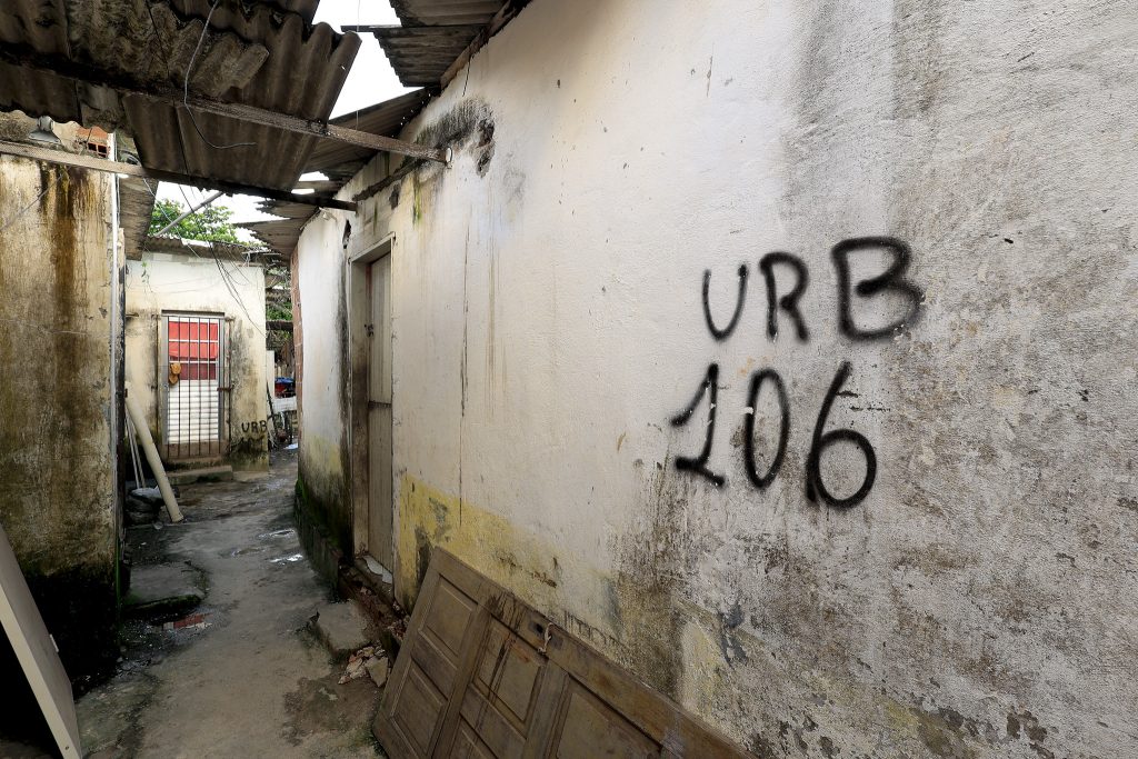 A foto mostra um beco estreito entre construções antigas e desgastadas. As paredes estão manchadas e com mofo, e há uma porta encostada no chão, sugerindo abandono ou reforma. No muro à direita, vê-se a inscrição “URB 106” feita com tinta preta em spray, enquanto ao fundo há outra marca semelhante. O telhado de zinco está danificado, com partes faltando e fios expostos, e o chão apresenta poças d’água e sujeira. A cena transmite uma atmosfera de degradação urbana, típica de áreas periféricas ou em processo de demolição.