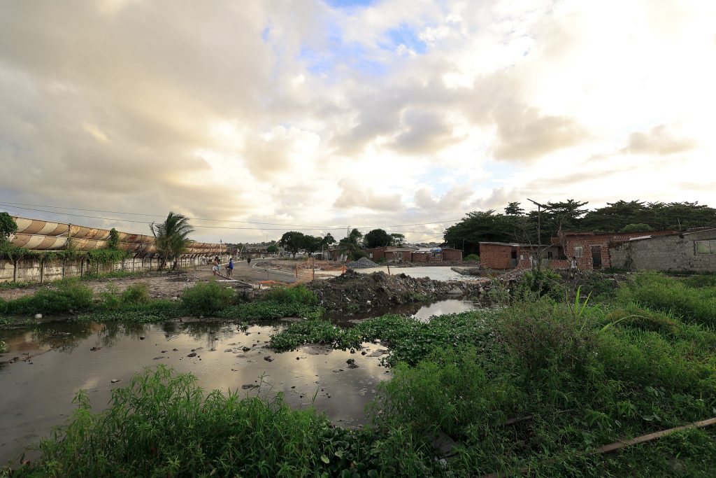 A imagem mostra uma área periférica em processo de transformação, com casas simples de tijolo e vegetação densa ao redor de um canal de água parada. O solo parece irregular e há entulho e lama, sugerindo obras ou reconstrução. À esquerda, vê-se uma estrutura metálica enferrujada coberta por plantas, e algumas pessoas caminhando ao longe. O céu está parcialmente nublado, com tons dourados e azulados, indicando o fim de tarde. A cena transmite uma mistura de vulnerabilidade ambiental e cotidiano urbano, onde a natureza e a ocupação humana coexistem em condições precárias.