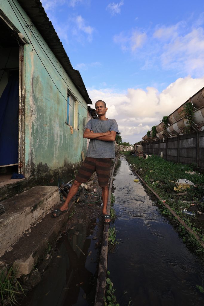 A foto mostra Alexsandro Silva em pé sobre uma calçada estreita ao lado de uma casa com paredes desgastadas e pintura verde-azulada descascada. Ele está com os braços cruzados, vestindo camiseta cinza e bermuda listrada, e olha firme para a câmera. Ao lado dele corre um canal estreito de água, cercado por vegetação e entulho, com uma muralha alta e enferrujada do outro lado. O céu azul com nuvens claras contrasta com o cenário urbano precário.