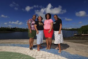 Quatro mulheres negras líderes da Poupança Comunitária da Ilha de Deus posam à beira do rio, com o manguezal e o skyline do Recife ao fundo. A foto foi tirada no calçadão da comunidade, com o horizonte urbano contrastando com a paisagem ribeirinha que define o território onde vivem e trabalham.