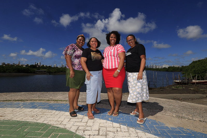 Quatro mulheres negras líderes da Poupança Comunitária da Ilha de Deus posam à beira do rio, com o manguezal e o skyline do Recife ao fundo. A foto foi tirada no calçadão da comunidade, com o horizonte urbano contrastando com a paisagem ribeirinha que define o território onde vivem e trabalham.