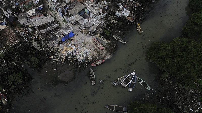A imagem mostra uma vista aérea de uma comunidade pobre à beira de um córrego escuro, cercada por casas improvisadas feitas de madeira e metal. Há entulho e lixo espalhados perto das moradias, e vários barcos pequenos de cores diferentes flutuam ou estão encostados na margem. Do outro lado do córrego, uma faixa de manguezal verde contrasta com o cenário urbano degradado, revelando a convivência entre natureza e precariedade.