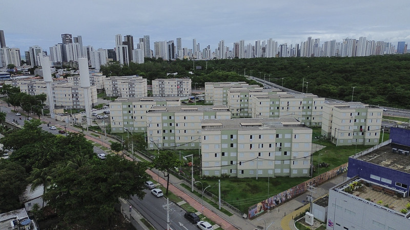 Vista aérea do conjunto habitacional Encanta Moça, no bairro do Pina, Zona Sul do Recife. Em primeiro plano, os blocos de quatro andares do habitacional contrastam com os prédios de alto padrão ao fundo — evidência visual da valorização imobiliária do entorno.