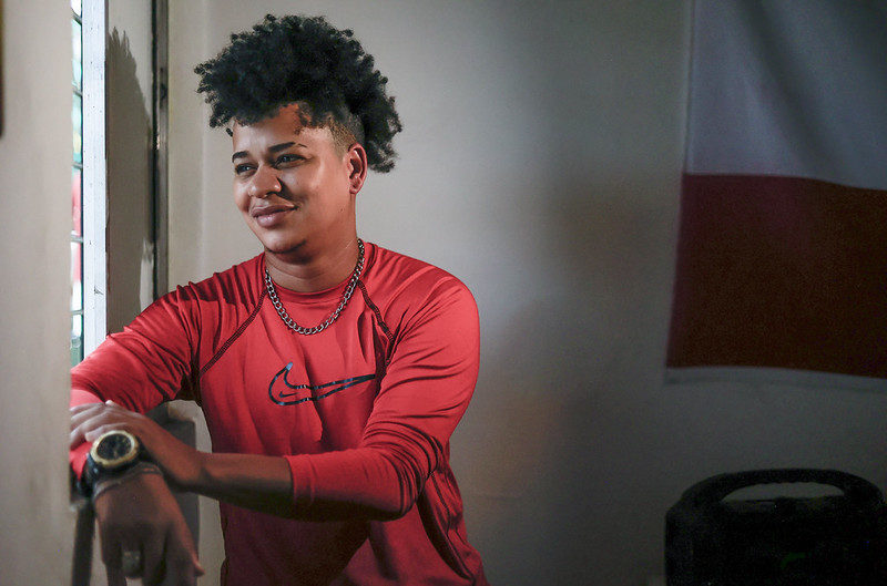 A imagem mostra uma pessoa jovem sentada dentro de um cômodo, próxima a uma janela por onde entra luz natural. Essa pessoa tem pele morena, cabelo curto e bem volumoso no topo, com as laterais raspadas. Está sorrindo levemente e olhando para fora da janela, com expressão tranquila. Veste uma camisa de manga comprida vermelha com o logotipo da Nike no peito, usa um relógio grande no pulso esquerdo e uma corrente prateada no pescoço. Ao fundo, há uma parede clara e parte de uma bandeira com duas faixas — branca em cima e vermelha embaixo — além de um objeto escuro que parece ser uma caixa de som. A luz suave destaca o rosto e o vermelho da roupa.
