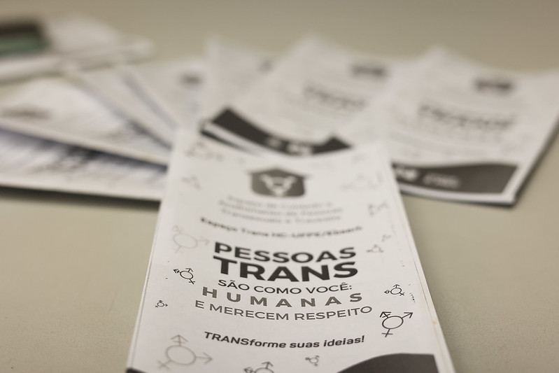 A imagem mostra alguns folhetos impressos dispostos sobre uma superfície clara, como uma mesa. O foco está em um folheto em primeiro plano, enquanto os outros aparecem desfocados ao fundo. O folheto principal é de cor branca e contém texto em português com mensagens de conscientização sobre respeito às pessoas trans. No centro, lê-se em letras grandes: “PESSOAS TRANS SÃO COMO VOCÊ: HUMANAS E MERECEM RESPEITO”. Abaixo, há a frase “TRANSforme suas ideias!”. O design inclui pequenos símbolos de gênero — masculino, feminino e trans — espalhados ao redor do texto. Na parte superior, há um logotipo de uma instituição, mas os detalhes não estão totalmente legíveis.