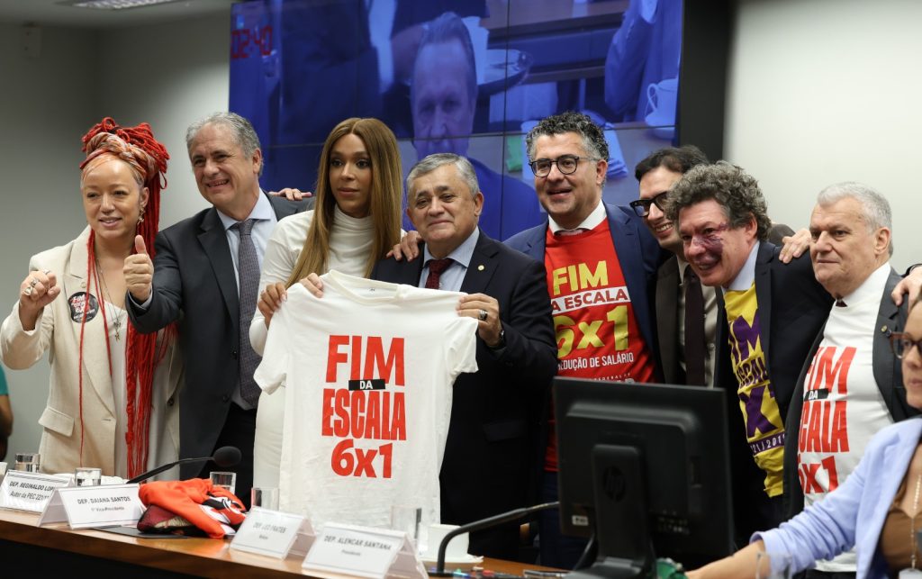 A foto mostra um grupo de pessoas em uma sala de reunião institucional, posando para uma foto coletiva. No centro, algumas delas seguram uma camiseta branca com a frase “FIM DA ESCALA 6x1”, enquanto outras vestem camisetas com a mesma mensagem. O ambiente parece ser uma audiência ou evento político, com mesas, microfones e um painel de vídeo ao fundo. As pessoas demonstram apoio à campanha, sorrindo e reunidas em torno da pauta apresentada.