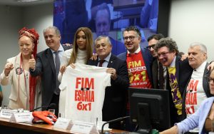 A foto mostra um grupo de pessoas em uma sala de reunião institucional, posando para uma foto coletiva. No centro, algumas delas seguram uma camiseta branca com a frase “FIM DA ESCALA 6x1”, enquanto outras vestem camisetas com a mesma mensagem. O ambiente parece ser uma audiência ou evento político, com mesas, microfones e um painel de vídeo ao fundo. As pessoas demonstram apoio à campanha, sorrindo e reunidas em torno da pauta apresentada.