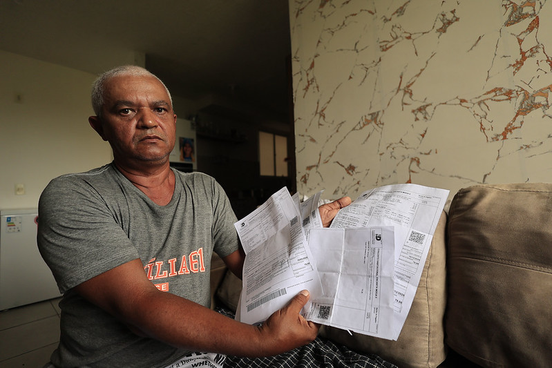 A imagem mostra Givaldo Rodrigues, um homem de meia-idade com cabelos grisalhos, sentado em um sofá dentro de casa. Ele segura várias contas ou boletos abertos nas mãos e tem uma expressão séria, sugerindo preocupação. Veste uma camiseta cinza com a palavra “VILLAGE” estampada e está em um ambiente simples, com parede clara de textura marmorizada ao fundo. A luz natural ilumina parte do rosto e do ambiente, revelando uma cena que transmite reflexão sobre dificuldades financeiras ou organização doméstica.